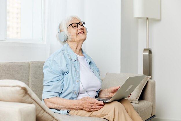 una mujer mayor emocional feliz con cabello gris está sentada en casa en un sofá acogedor con auriculares en la cabeza y sonriendo felizmente sosteniendo una computadora portátil en el regazo
