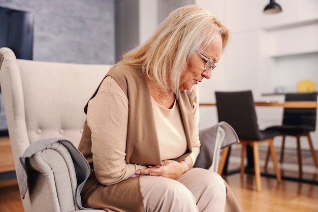 Mujer mayor enferma sentada en su casa en su silla y sosteniendo su estómago