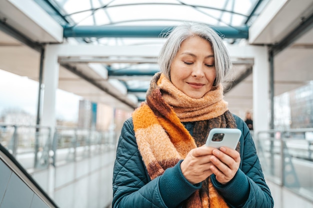 Mujer mayor envía un mensaje con un teléfono móvil en una estación de transporte