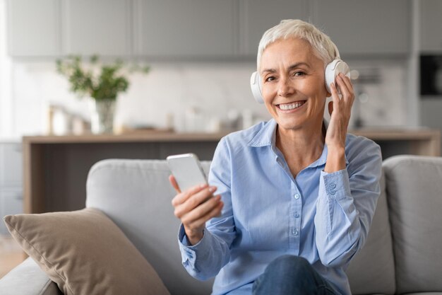 La mujer mayor escucha música con auriculares y teléfono en casa