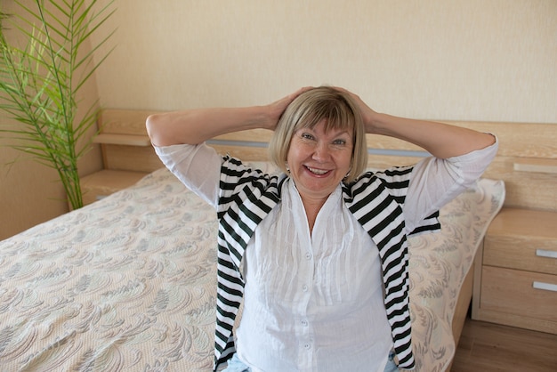 Mujer mayor feliz con cabello gris relajante sonriendo está mirando a la cámara en su casa en el dormitorio.