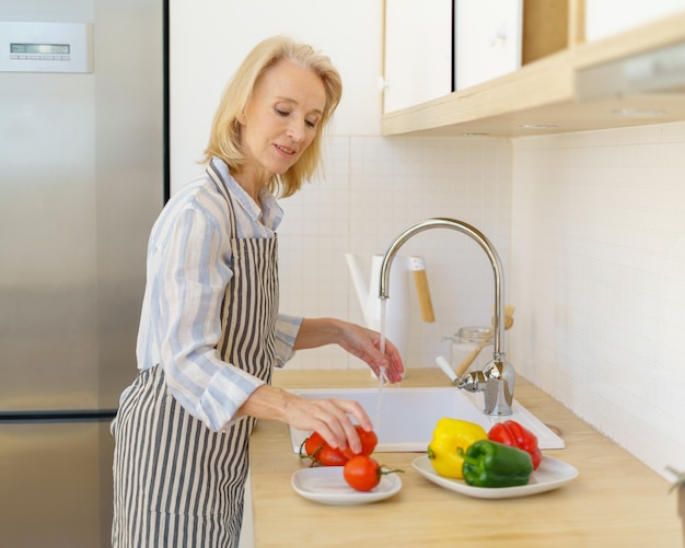 Mujer mayor feliz en delantal preparando verduras para ensalada mientras se cocina en la cocina moderna en casa
