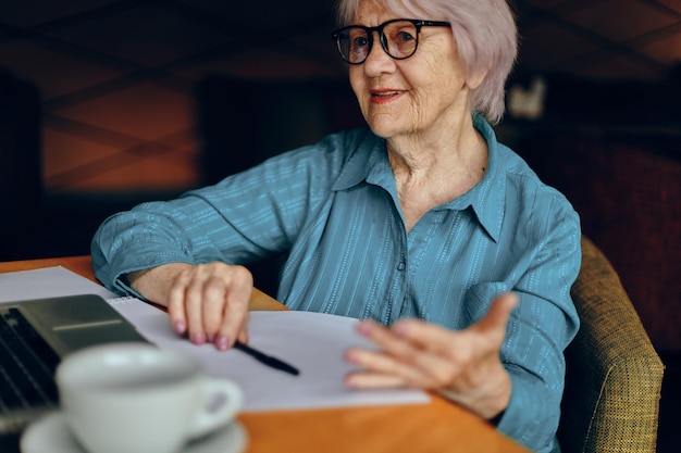 Mujer mayor feliz sentada en un café con una taza de café y una computadora portátil Mujer jubilada charlando inalterada