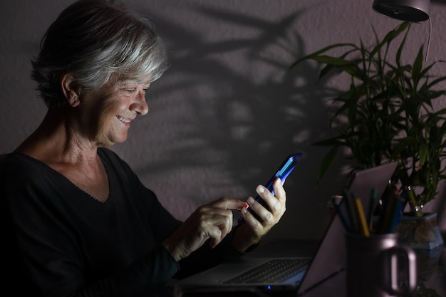 Mujer mayor feliz trabajando y usando la computadora portátil a altas horas de la noche, mirando el mensaje del teléfono móvil