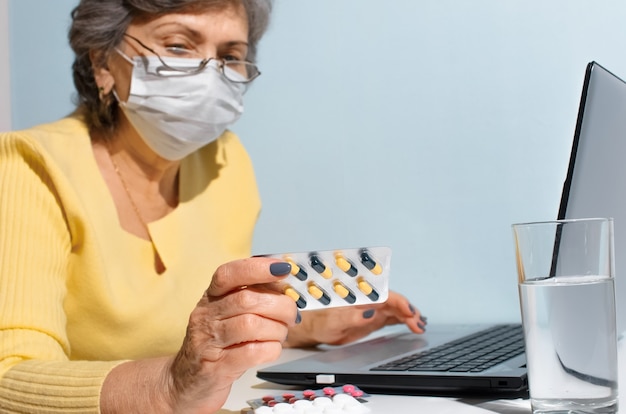 Mujer mayor con gafas y máscara médica leyendo instrucciones de medicina, enfoque selectivo. Anciana usando laptop en casa. Concepto de consulta en línea con el médico, nueva normalidad.