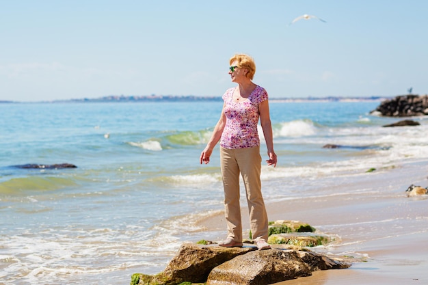 Mujer mayor con gafas de sol se para en la playa y mira al mar