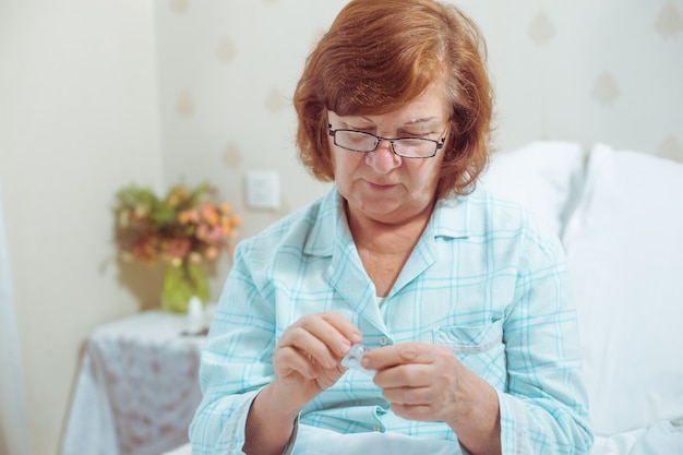 Mujer mayor en glassessitting en la cama en casa tomando pastillas Abuela de pelo rojo caucásico enfermo