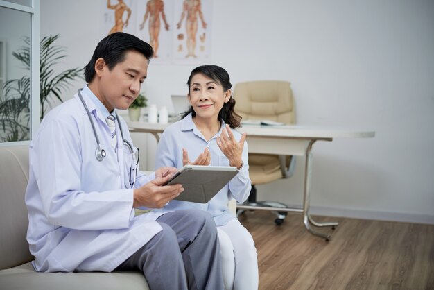 Mujer mayor hablando con el médico de cabecera sobre los síntomas de la enfermedad cuando él está revisando su historial médico en tableta digital