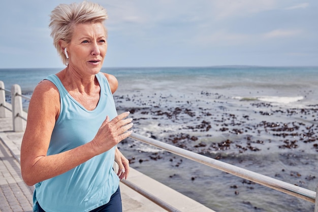 Mujer mayor haciendo ejercicio y corriendo en la maqueta del cielo del paseo marítimo o energía de la salud, bienestar y entrenamiento Auriculares femeninos mayores y fitness en el océano de deportes, corredor de cardio y mentalidad fuerte