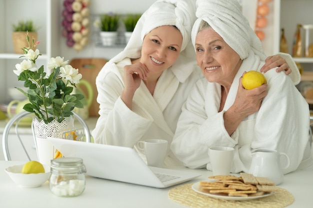 Foto mujer mayor, y, hija, en, un, albornoz, con, computadora portátil, y, té