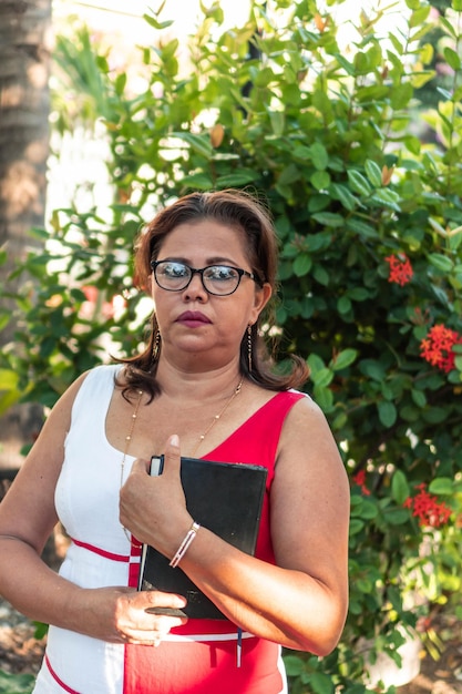 Mujer mayor hispana sosteniendo un libro en sus manos caminando en el parque. Profesor con ropa elegante y gafas.