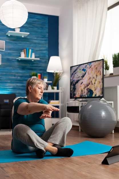 Mujer mayor jubilada en ropa deportiva sentada en la estera de yoga viendo la lección de fitness en la computadora portátil practicando ejercicios de brazo para adelgazar peso