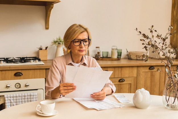 Mujer mayor madura de mediana edad con factura en papel o carta en casa para realizar pagos en línea en el sitio web, calcular el costo de la tarifa de impuestos financieros, revisar la cuenta bancaria.