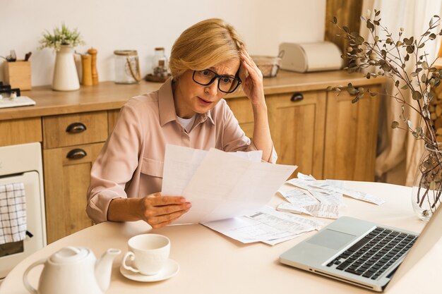 Mujer mayor madura de mediana edad con factura en papel o carta usando una computadora portátil en casa para realizar pagos en línea en el sitio web, calcular el costo de la tarifa de impuestos financieros, revisar la cuenta bancaria.