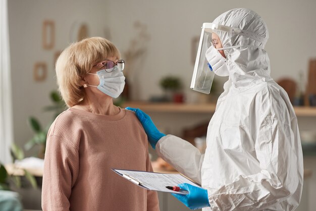 Mujer mayor en máscara consultando con médico en traje protector durante su visita