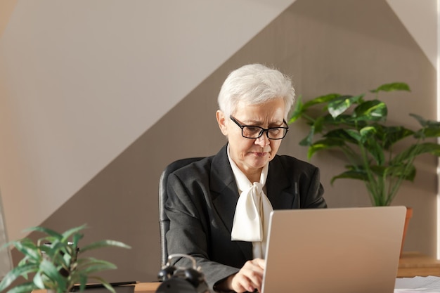 Mujer mayor de mediana edad europea con estilo segura que usa una computadora portátil en el lugar de trabajo Mujer de negocios de cabello gris de los años 60 madura y elegante sentada en la mesa de la oficina Trabajadora profesional de maestro líder jefe