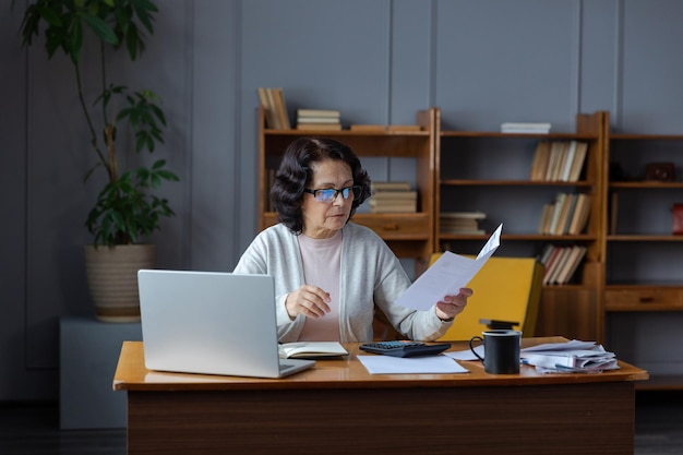 Mujer mayor de mediana edad sentada con una computadora portátil y un documento en papel pensativa señora madura mayor leyendo papel