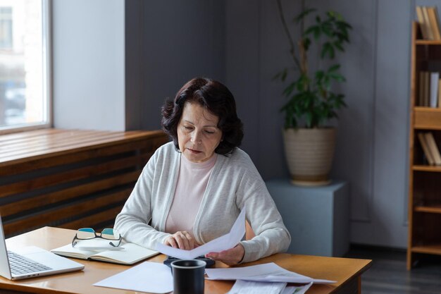 Mujer mayor de mediana edad sentada con una computadora portátil y un documento en papel pensativa señora madura mayor leyendo papel
