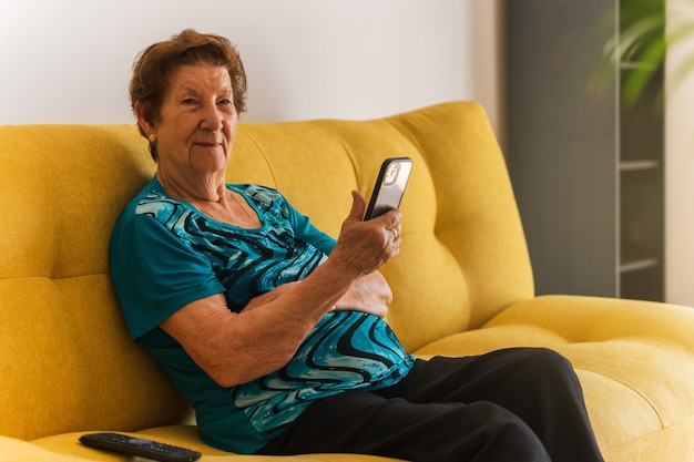 Foto mujer mayor mirando a la cámara a través de un teléfono móvil mientras se sienta en el sofá