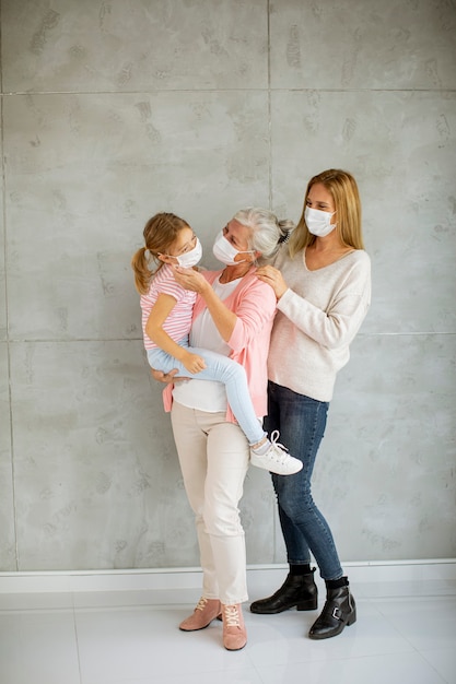 Mujer mayor, mujer adulta y niña linda, tres generaciones con máscaras faciales protectoras en casa