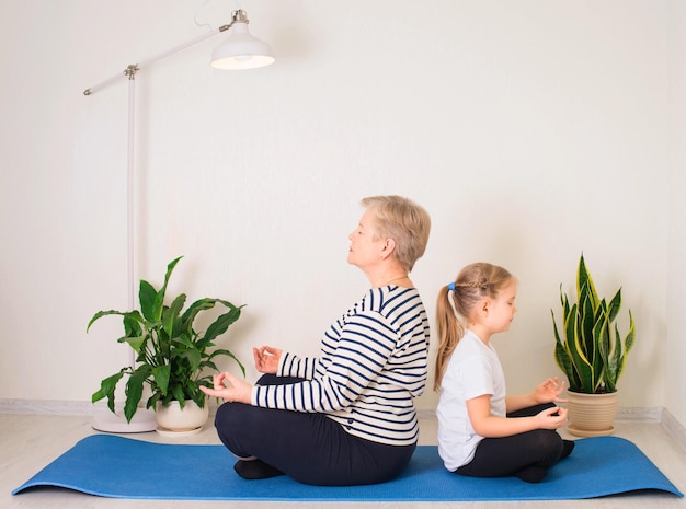 Mujer mayor y niño meditando en casa en posición de yoga espalda con espalda en colchoneta de fitness