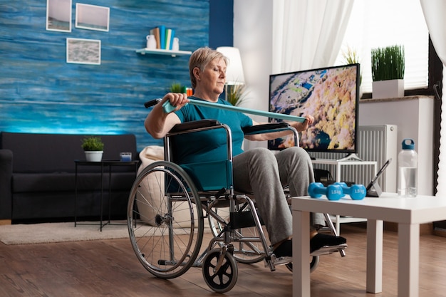 Foto mujer mayor no válida en silla de ruedas con banda elástica de resistencia que estira los músculos del cuerpo recuperándose después de un accidente de discapacidad viendo videos de entrenamiento en tableta