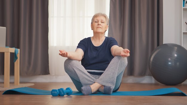 Mujer mayor pacífica con los ojos cerrados haciendo yoga en la sala de estar. Activo estilo de vida saludable deportivo anciano entrenamiento entrenamiento bienestar en el hogar y ejercicio en interiores