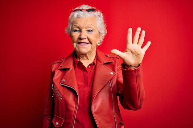 Mujer mayor de pelo gris con chaqueta roja informal y gafas de sol mostrando y señalando con los dedos número cinco mientras sonríe confiada y feliz