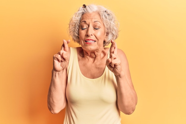 Mujer mayor de pelo gris con ropa informal haciendo gestos con los dedos cruzados sonriendo con esperanza y los ojos cerrados suerte y concepto supersticioso