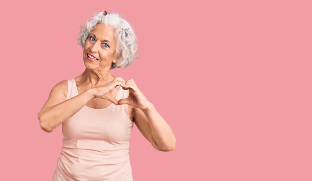 Mujer mayor de pelo gris con ropa informal sonriendo enamorada mostrando el símbolo del corazón y la forma con las manos concepto romántico