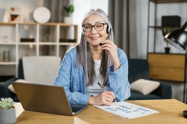 Mujer mayor positiva que usa auriculares y una computadora portátil para una conversación de video en la oficina