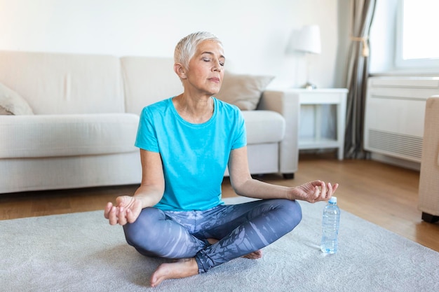 Mujer mayor practicando yoga sentada en el ejercicio Half Lotus Ardha Padmasana plantean trabajando usando ropa deportiva sesión de meditación interior interior de casa de cuerpo entero