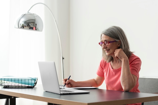 Mujer mayor que estudia en casa mientras usa la computadora portátil y toma notas