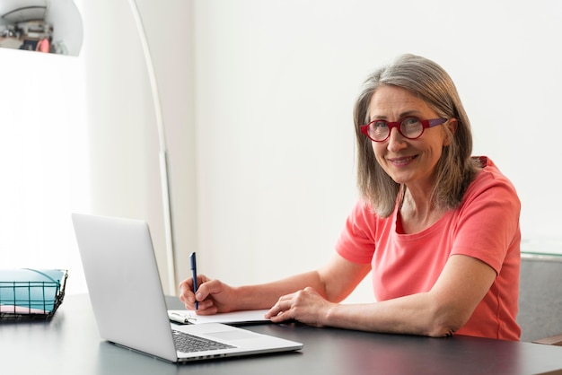 Mujer mayor que estudia en casa mientras usa la computadora portátil y toma notas