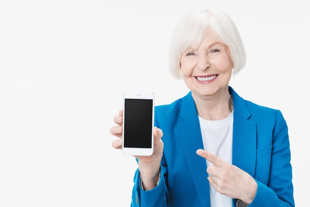 Mujer mayor que muestra la pantalla del teléfono inteligente sobre un fondo aislado que señala con el dedo