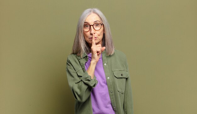 Mujer mayor que parece seria y cruzada con el dedo presionado a los labios exigiendo silencio o silencio, manteniendo un secreto