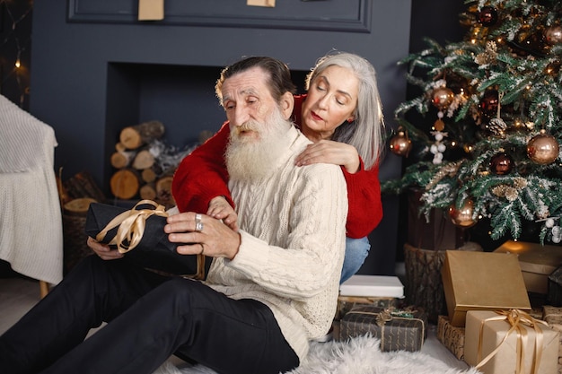 Mujer mayor que presenta un regalo de navidad a su esposo sentado cerca del árbol de navidad