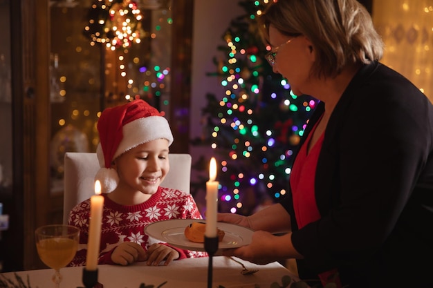 Mujer mayor que sirve el plato de comida al niño sonriente. Familia feliz cenando en casa de Navidad cerca del árbol de Navidad decorado.