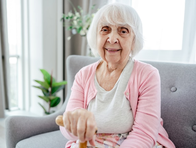 Mujer mayor que sostiene el bastón, sentado y mirando hacia atrás sonriendo. Retrato de anciano pensionista en casa