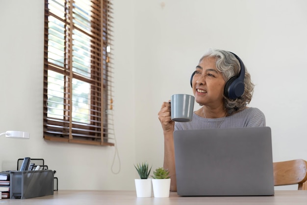 Mujer mayor que usa una computadora portátil mientras usa auriculares en casa Concepto de estilo de vida y tecnología para personas mayores alegres
