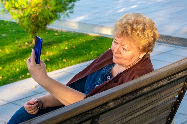 Mujer mayor que usa el teléfono inteligente, lee libros electrónicos, leasten música o toma educación en línea en el parque en un banco
