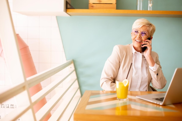 Mujer mayor que usa teléfono móvil mientras trabaja en una computadora portátil y bebe jugo de naranja fresco en el café