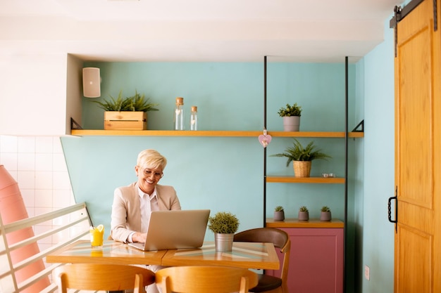 Mujer mayor que usa teléfono móvil mientras trabaja en una computadora portátil y bebe jugo de naranja fresco en el café