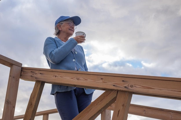 Mujer mayor relajada con gorra y anteojos de pie al aire libre al amanecer sosteniendo una taza de café