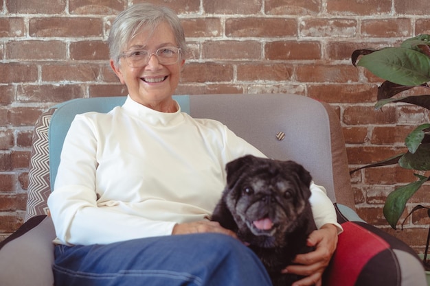 Mujer mayor relajada sonriendo sentada en casa en un sillón cerca de su amor de perro pug y el concepto de mejor amiga