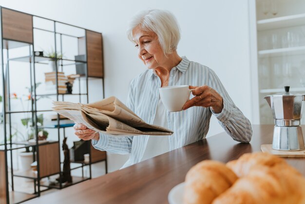 Mujer mayor relajándose en casa, leyendo el periódico y desayunando