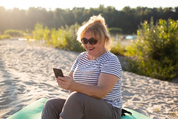 Mujer mayor sentada en una arena y con smartphone en la playa de verano