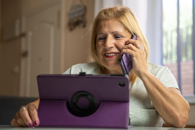 Mujer mayor sentada en casa mientras habla por su teléfono celular con otra persona y se entretiene con su tableta
