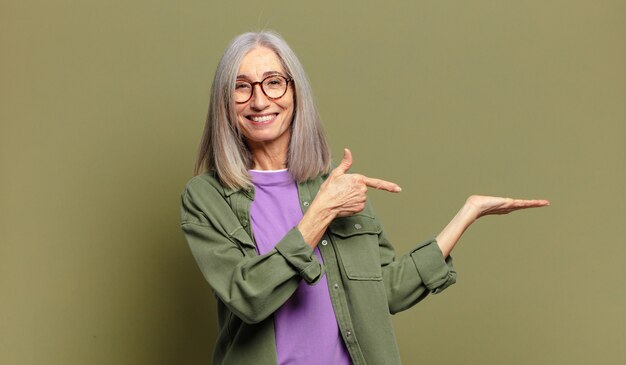 Foto mujer mayor sonriendo, sintiéndose feliz, despreocupada y satisfecha, apuntando al concepto o idea en el espacio de la copia en el lateral