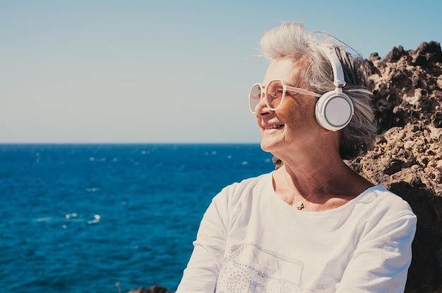 Mujer mayor sonriente y despreocupada escuchando música con auriculares sentada y mirando el horizonte sobre el mar